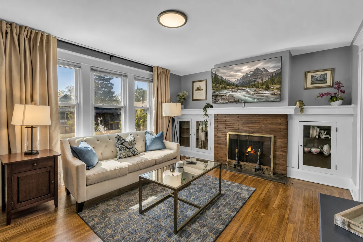 Furnished living room with a lit fireplace, cream sofa with blue cushions, and hardwood floors